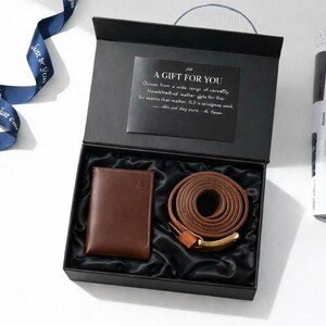 May include: A gift set in a black box, featuring a brown leather wallet and belt with a gold-colored buckle. The box contains a card with text that reads "A GIFT FOR YOU". A blue ribbon with the text "Just For You" is visible.