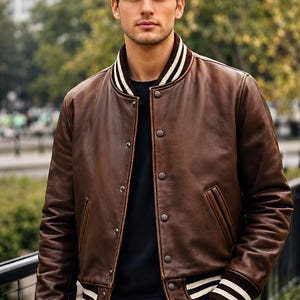 Peut inclure: Un blouson style teddy en cuir marron doté d'une fermeture à boutons-pression, de deux poches latérales, ainsi que de poignets et d'un ourlet côtelés à rayures blanches et marron. Il est porté sur un haut sombre et un jean bleu classique.