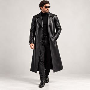 May include: A long black leather trench coat featuring a wide collar, button-front closure, and long sleeves. The coat is worn over a black turtleneck sweater and black trousers, paired with black leather boots and dark-tinted sunglasses.