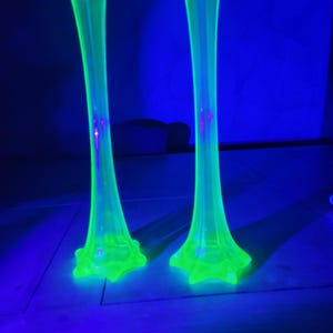 May include: Two tall, slender, neon green glass vases with flared bases and tops. The vases are illuminated with a black light, creating a glowing effect against a dark blue background. The vases are likely vintage or antique.