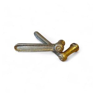 Op de afbeelding: Twee antieke deurklinken. Eén klink is zilverkleurig met een gestippelde rand en bloemaccenten. De andere klink is goudkleurig met een vergelijkbaar bloemmotief. De klinken zijn waarschijnlijk van metaal.