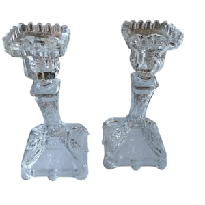 May include: Two clear glass candle holders. Each has a square base, a curved stem, and a scalloped top. The glass is intricately detailed with floral patterns. The candle holders are designed to hold a single candle.