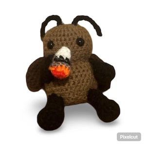 Peut inclure: Un hibou en crochet marron fait à la main avec des yeux, des ailes et des pieds noirs. Le hibou tient un petit objet coloré avec des détails blancs, orange et noirs. Le hibou a deux antennes noires et est posé sur un fond blanc.