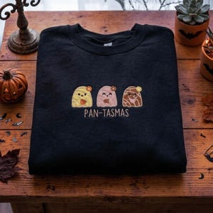 May include: A black crewneck sweatshirt featuring an embroidered design of three ghost-like figures resembling pan dulce, each with a small flower on top. Below the figures, the text PAN-TASMAS is embroidered in a light beige thread.
