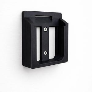 May include: A black wall-mounted bracket with a rectangular design. Two screws secure the bracket to the wall. The bracket has a slot for holding an item. The item is mounted on a white wall.