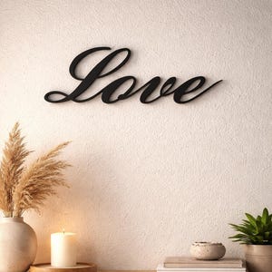 Peut inclure: Un signe mural noir en forme de mot "Love" en écriture cursive est fixé sur un mur blanc texturé. En dessous, un meuble en bois présente des éléments décoratifs.