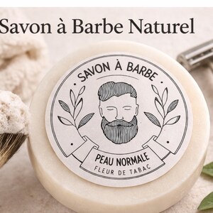 Pode incluir: Uma barra redonda de sabonete natural para barba, de cor esbranquiçada, com um rótulo a preto e branco com o rosto de um homem barbudo. O texto "Savon à Barbe Naturel" está no topo. Uma escova de barbear, uma navalha e uma toalha também estão na imagem.