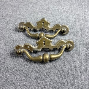 以下が含まれることがあります： 鍵穴のディテールが施された、真鍮製のアンティーク引き出し取っ手2つ。取っ手は曲線的なハンドルデザインと装飾的なアクセントが特徴です。真鍮は暖かく、古びた風合いで、ヴィンテージまたはレトロなスタイルを連想させます。