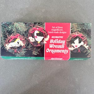 May include: A boxed set of three decorative holiday wreath ornaments. Each wreath features a different design: a dog, a goose, and a dove. The wreaths are made of twigs and decorated with red bows and white flowers. The box has text that reads "Holiday Wreath Ornaments."
