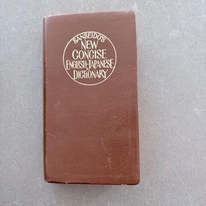 Peut inclure: Un livre rectangulaire marron avec le titre "Sanseido's New Concise English-Japanese Dictionary" dans un design circulaire blanc. Le livre a une apparence usée.