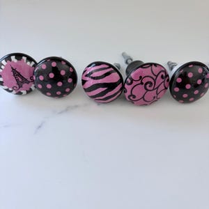 May include: Five round cabinet knobs with metal hardware. The designs include a black Eiffel Tower on a pink background, black and pink polka dots, pink and black zebra stripes, black swirls on a pink background, and black and pink polka dots.