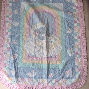 Pode incluir: Uma colcha de bebê em tons pastel com uma imagem central de um bebê dormindo sob um arco-íris. A colcha apresenta um fundo azul com padrões florais e geométricos, uma borda rosa com babados e provavelmente é feita de tecido macio.