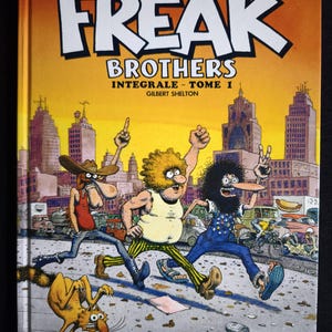 Op de afbeelding: Stripboekcover voor "Les Fabuleux Freak Brothers - Intégrale - Tome 1" van Gilbert Shelton. De cover toont drie stripfiguren die rennen voor een skyline. De titel staat in grote witte letters.
