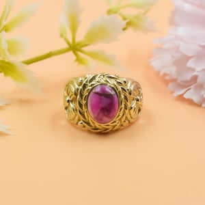 May include: A gold-colored ring with an oval, magenta-colored stone. The ring has an ornate, floral design around the stone. The background is a light peach color with white and pink flowers.