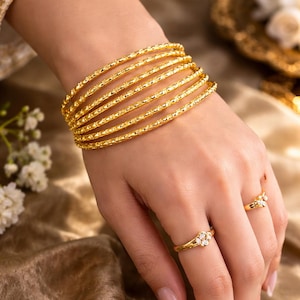 Może przedstawiać: Zestaw złotych bransoletek i dwa złote pierścionki z małymi białymi kamieniami. Bransoletki są ułożone na nadgarstku, a pierścionki na palcach. Biżuteria jest umieszczona na złotym tle z tkaniny.