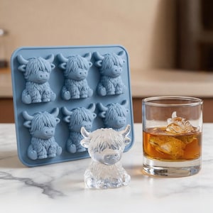 May include: A blue silicone ice cube tray with six Highland cow-shaped molds. A clear ice sculpture of a Highland cow sits in front of the tray. A glass of amber liquid contains a Highland cow ice cube.