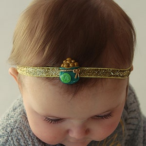 May include: A baby wearing a light blue sweater and a gold headband with a green pot of gold charm. The headband is decorated with glitter.