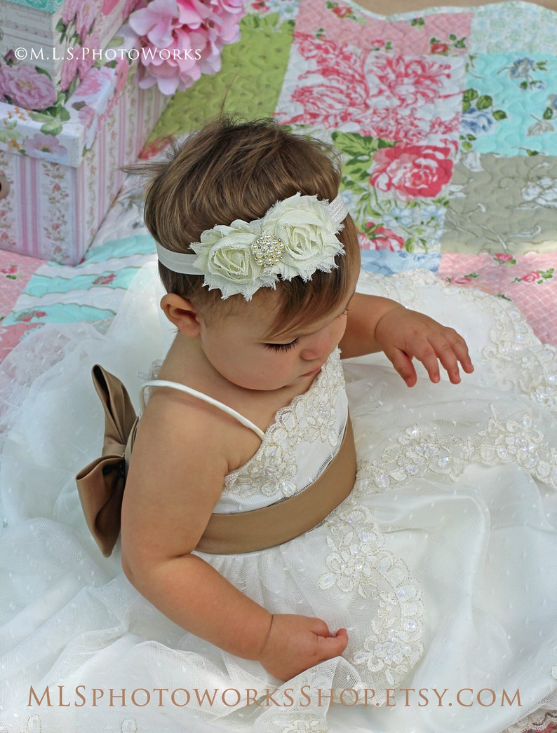 May include: A baby girl wearing a white dress with lace trim and a brown sash. She is wearing a white flower headband with a rhinestone accent. The baby is sitting on a floral patterned quilt.