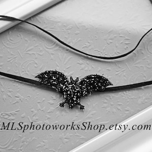 May include: A black bat-shaped headband with glitter accents. The headband is on a white background with a floral pattern.
