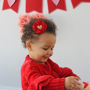 Peut inclure: Une jeune fille aux cheveux bruns boucl&eacute;s porte un pull rouge et un barrette de cheveux en forme de fleur rouge avec un c&oelig;ur rose au centre. Elle joue avec un jouet jaune.