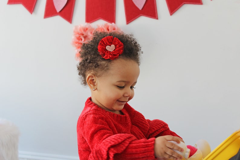 Peut inclure: Une jeune fille aux cheveux bruns boucl&eacute;s porte un pull rouge et un barrette de cheveux en forme de fleur rouge avec un c&oelig;ur rose au centre. Elle joue avec un jouet jaune.