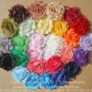 May include: A collection of 20 handmade fabric flowers in various colors, including brown, green, light green, yellow, orange, red, pink, white, blue, and purple. The flowers are arranged in a circle on a brown surface.