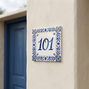 Könnte beinhalten: Ein quadratisches Hausnummernschild aus Keramik mit der Ziffernfolge 101 in dunkelblauer Schreibschrift. Der Rand ist mit einem handgemalten Muster aus blauen Blättern, kleinen Granatäpfeln und Vögeln auf weißem Grund mit Goldakzenten verziert.
