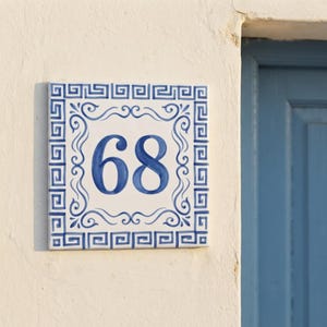Op de afbeelding: Een vierkante keramische huisnummertegel met het nummer 68 in blauwe verf. De tegel heeft een witte achtergrond met een blauw Grieks sleutelpatroon als rand en decoratieve blauwe krullen rondom de centrale cijfers op het oppervlak.