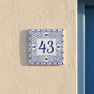 Könnte beinhalten: Ein quadratisches Hausnummernschild aus Keramik mit der blauen Ziffer 43. Das Design zeigt ein blaues Zickzack-Muster mit kleinen blauen Fischsymbolen auf weißem Grund, eingerahmt von einer blau-weißen geometrischen Bordüre.