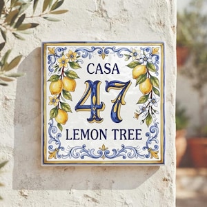 Może przedstawiać: Kwadratowa ceramiczna tabliczka z numerem domu z napisem CASA 47 LEMON TREE ciemnoniebieskimi literami. Wzór zawiera ręcznie malowane żółte cytryny, zielone liście i białe kwiaty, otoczone ozdobną niebiesko-złotą bordiurą z ornamentami.