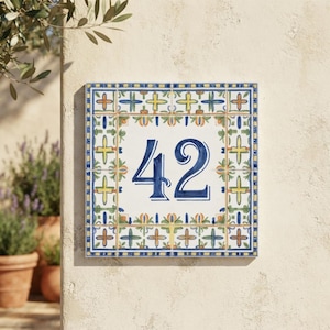 Può includere: Una targa quadrata in ceramica per numero civico che mostra le cifre 42 in un carattere blu audace. Il centro è bianco, circondato da un bordo decorativo con motivi floreali gialli, verdi e arancioni su sfondo bianco con finiture blu.