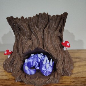 Op de afbeelding: Een bruine boomstamsculptuur met een uitgehold midden, met een paars drakenbeeldje. Twee rood-witte paddenstoelbeeldjes zijn bevestigd aan de boomstam. De draak is opgerold in de boom.