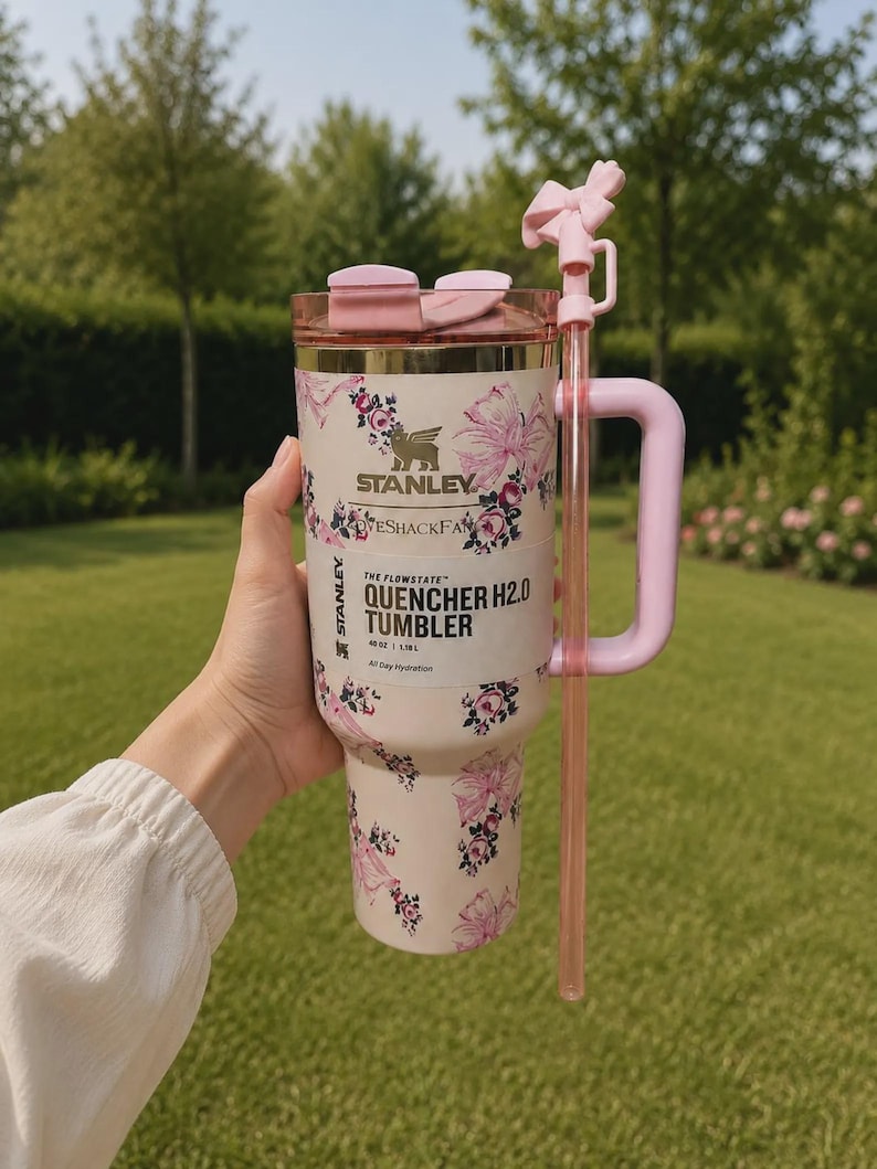 May include: A light pink Stanley Flowstate Quencher H2.0 tumbler with a floral pattern, featuring a pink handle, a matching lid, and a pink straw with a bow-shaped topper. The tumbler displays the Stanley and LoveShackFancy brand logos.