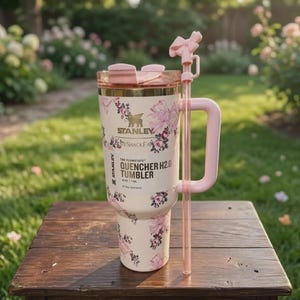May include: A cream-coloured Stanley Quencher H2.0 tumbler with a floral pattern, featuring a pink handle, a matching pink lid, and a clear straw topped with a pink bow accessory. The tumbler displays the text Stanley, LoveShackFancy, and 40 oz / 1.18 L.