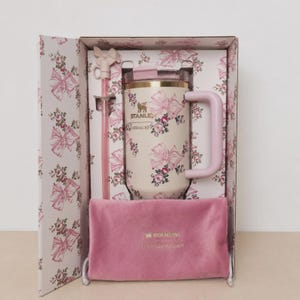 May include: A Stanley tumbler with a light pink floral and bow pattern, featuring a matching straw with a bow topper, a light pink handle, and a clear lid. The set is presented in a matching floral box with a soft pink velvet pouch with Stanley text.
