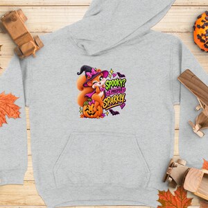 Op de afbeelding: Een lichtgrijze hoodie met een afbeelding van een cartoon-eekhoorn die een heksenhoed draagt en op een pompoen zit. De tekst "SPOOKY? I PREFER SPARKLY" staat in kleurrijke, gestileerde letters, omringd door kleine vleermuizen, sterren en hartjes.