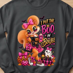 May include: A dark gray sweatshirt featuring a cartoon squirrel wearing a pink polka dot bow, sunglasses, and a black outfit with a pumpkin graphic. The text reads I PUT THE BOO IN BOUJEE, accompanied by a ghost, pumpkins, and bats in pink and black.