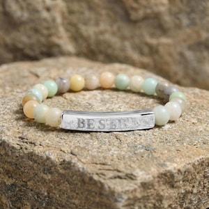 Può includere: Braccialetto con perline rotonde color pastello. Presenta una barra rettangolare color argento con la scritta incisa "BE STILL". Il braccialetto è appoggiato su una superficie rocciosa strutturata. Le perline sono verdi, gialle e pesca.