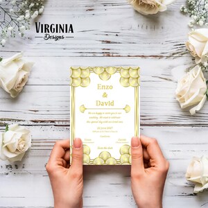 May include: A wedding invitation with gold Art Deco style design elements. The invitation features the names "Enzo & David" and the date "22 June 2027". The invitation is held by a person's hands, surrounded by white roses and greenery.