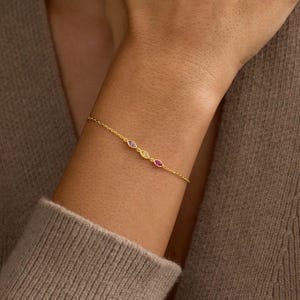 Könnte beinhalten: Ein zartes Goldkettenarmband mit drei kleinen, marquise-geschliffenen Edelsteinen in den Farben Hellviolett, Gelb und Dunkelrosa. Das Armband wird durch eine dünne, polierte Goldgliederkette gehalten, die auf der Haut des Handgelenks aufliegt.