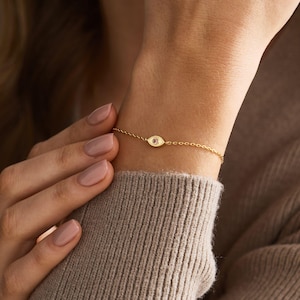 Op de afbeelding: Een verfijnde gouden kettingarmband met een centraal oogvormig bedeltje. Het bedeltje is ontworpen met een gouden omlijning en een klein steentje in het midden. De armband is gemaakt van fijne, onderling verbonden schakels om de pols.