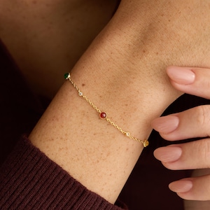 Könnte beinhalten: Ein zartes Goldkettenarmband mit kleinen, gleichmäßig verteilten runden Edelsteinen in Rot und Grün. Das Armband ist mit einem einfachen Verschluss versehen und liegt auf der Haut, was ein schlichtes Design mit farbigen Akzenten unterstreicht.
