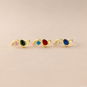 May include: Three gold rings, each featuring a small round gemstone and a larger pear-shaped gemstone. The first ring has a pink round stone and a green pear stone. The second has a blue round stone and a red pear stone. The third has a clear round stone and a blue pear stone.