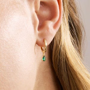 May include: A gold hoop earring featuring a small, teardrop-shaped green gemstone charm suspended from the bottom. The earring is worn in the earlobe, showcasing a polished metallic finish and a vibrant, deep green color on the stone pendant.