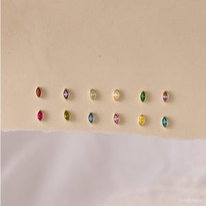 May include: Twelve individual stud earrings arranged in two rows. Each earring features a small, marquise-cut gemstone set in a gold-toned metal bezel. The stones display a variety of colors including red, purple, green, yellow, pink, and blue.