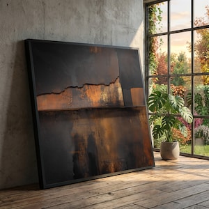 Puede incluir: Gran impresión artística abstracta enmarcada con una paleta de colores oscuros. La obra de arte presenta una superficie texturizada con tonos de negro, marrón y dorado. El arte enmarcado está apoyado contra una pared de hormigón, con una ventana y una planta en el fondo.