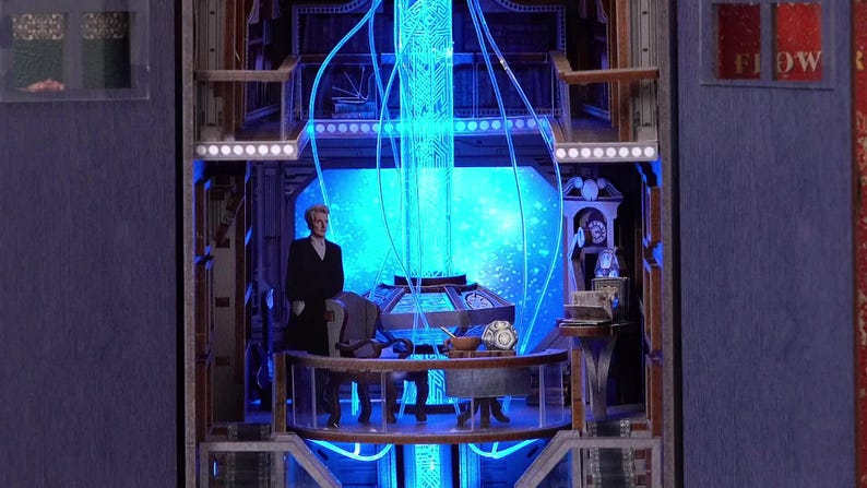 May include: A detailed diorama featuring a figure in a black coat standing in a futuristic interior with blue neon lighting. The scene includes a control panel, a starry backdrop, and the word "FLOW".