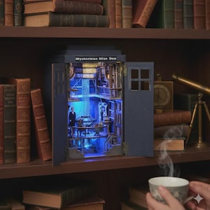 May include: A miniature blue box with the text "Mysterious Blue Box" on top, with open doors revealing a detailed interior with figures and blue lighting. The box is on a bookshelf surrounded by books.