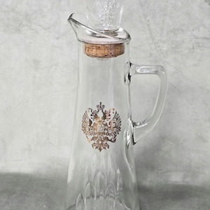 May include: A clear glass pitcher featuring a gold-colored double-headed eagle emblem on the front, a cork stopper, and a small glass cup resting on top. The pitcher has a handle, a pouring spout, and a textured base with vertical glass ridges.