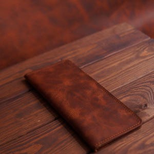 May include: A brown leather wallet with a textured surface rests on a wooden surface. The wallet is rectangular and stitched along the edges. The wood has a dark brown colour with visible grain.